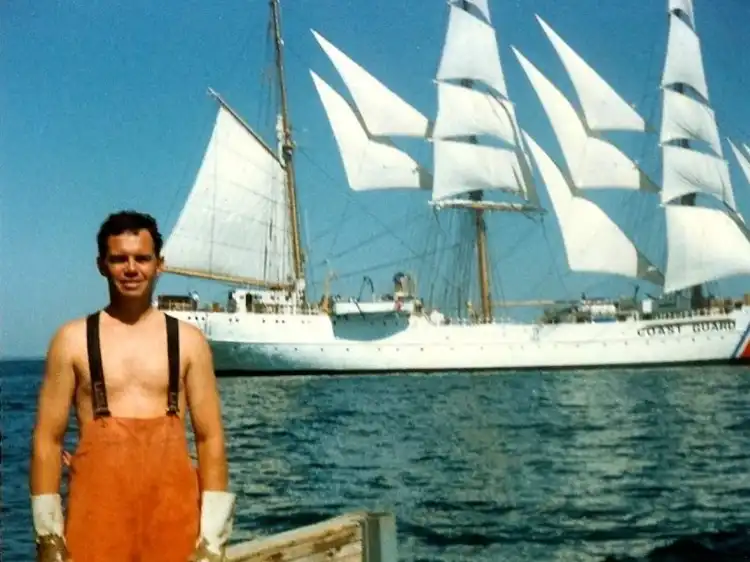Man in orange overalls standing by a dock with a large white sailing ship in the background.