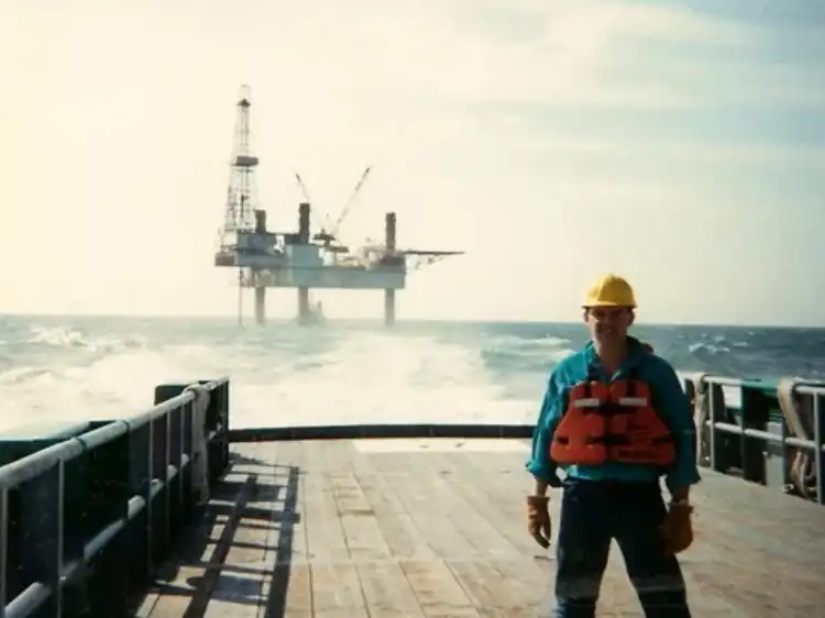 Worker in safety gear on offshore oil platform deck with rig in background.