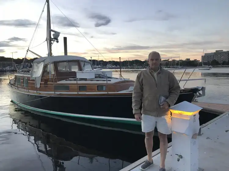 Man standing on a dock beside a classic wooden boat at sunset.