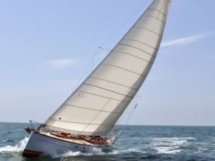 A sailboat with a large white sail glides on the ocean under clear skies.