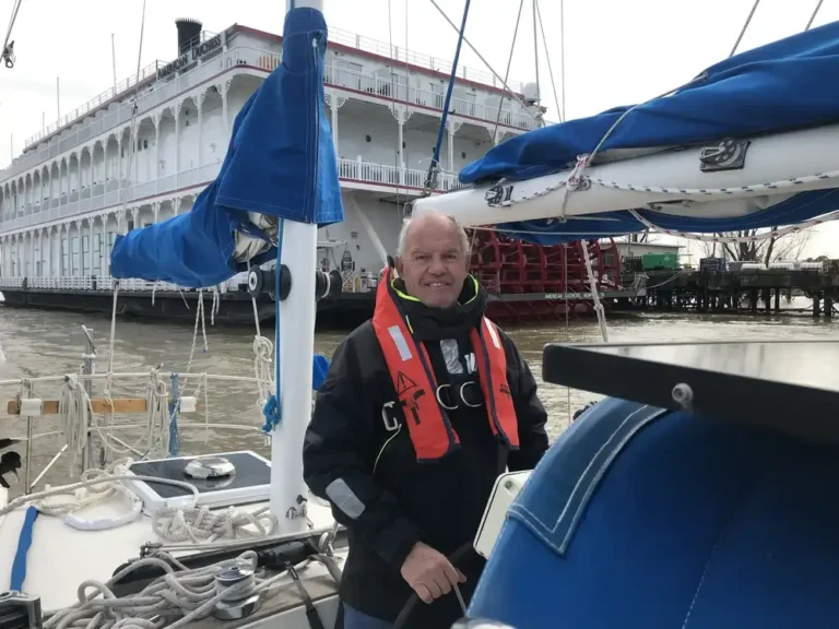 Man in sailing gear standing on a boat near a docked ship.