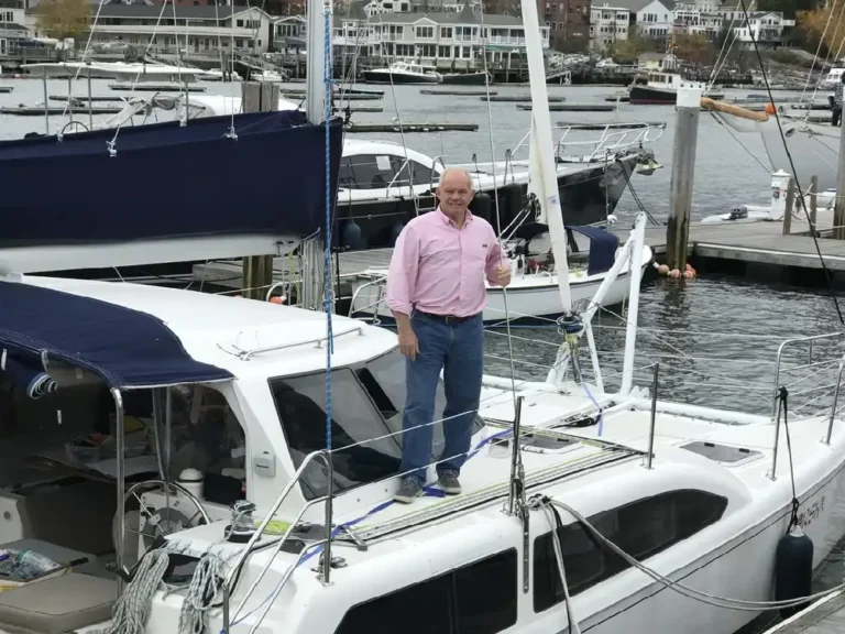 Man standing on a docked white sailboat in a marina.