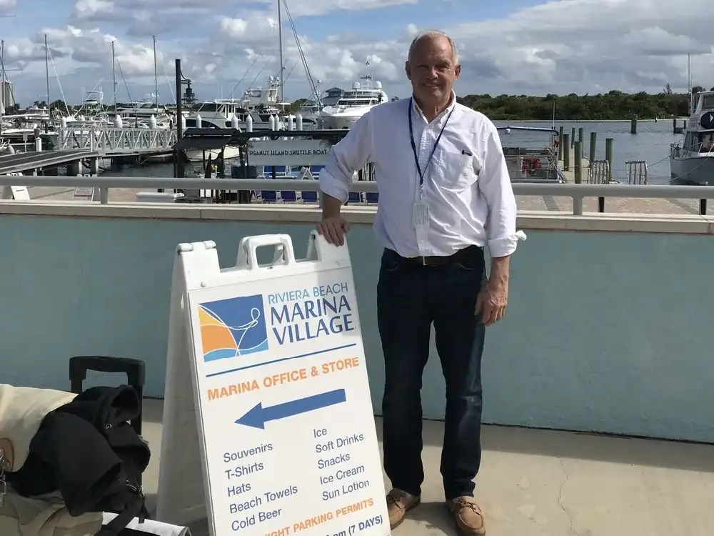 Man standing near marina sign with boats in the background.
