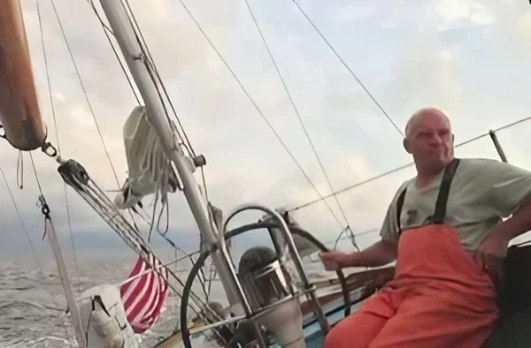 Man steering a sailboat wearing orange overalls with an American flag.