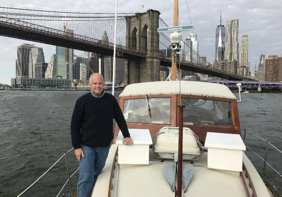 Man standing on a boat with Brooklyn Bridge and NYC skyline in the background.
