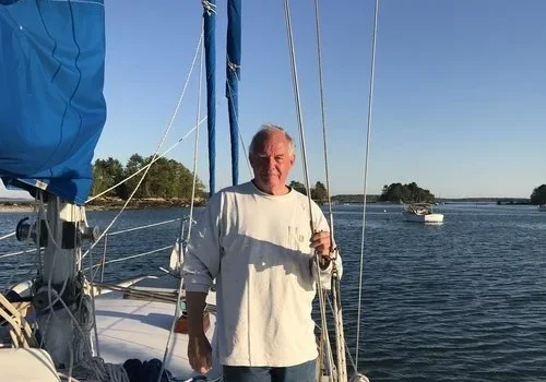 Man standing on a sailboat holding a drink with water and islands in the background.