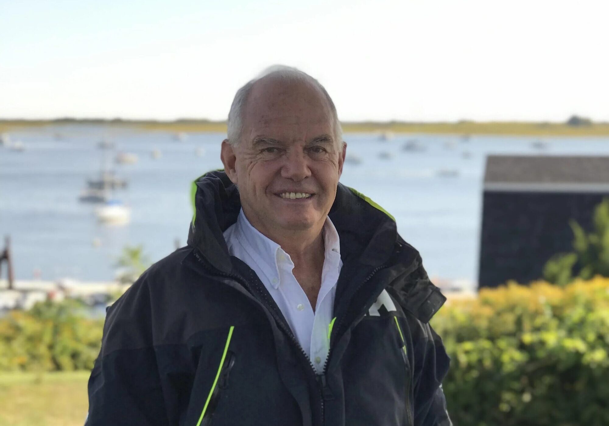 Smiling man outdoors near a body of water with boats in the background.
