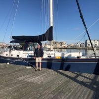 Person standing on dock beside a sailboat with cityscape in background.