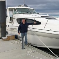 Man posing cheerfully on a dock beside a large white yacht.