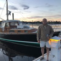 Man standing on dock near a classic wooden sailboat at sunset.