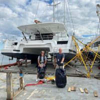 Two people posing in front of a large catamaran docked at a marina.